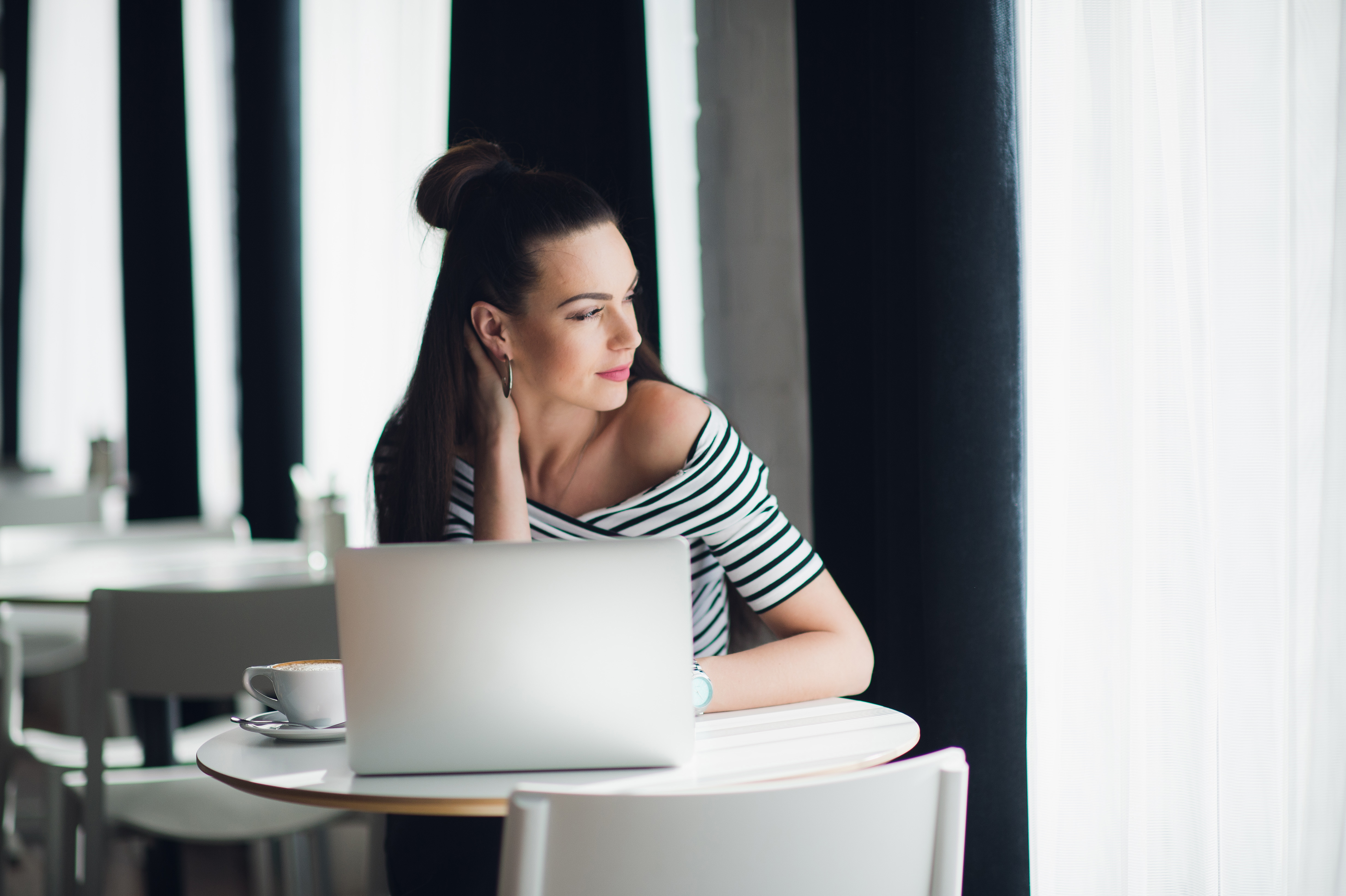 Smiling female writer is working with her new idea at lunch and looking through the window for searching for inspiration. Attractive adult woman sitting at the table with a cup of coffee and a laptop.