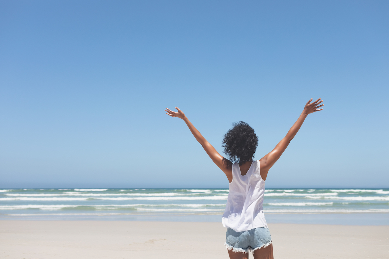 Rear view of beautiful young mixed-race woman standing with open arm at beach on a sunny day. she seems happy